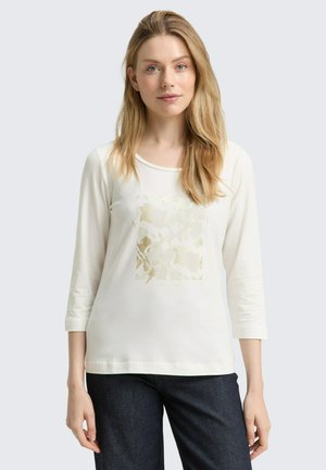 Blonde Frau in einem weißen Shirt mit drei Viertel Ärmeln und einem dezenten beigen abstrakten Muster, dazu eine dunkelblaue Jeans, stehend vor einem hellen Hintergrund.
