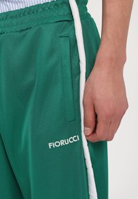 Žalios sportinės kelnės su lygia tekstūra, su baltomis šoninėmis juostomis ir balta "Fiorucci" logotipu, sukurta su šoniniu kišenė.