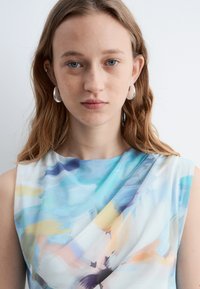 Jeune femme aux cheveux châtain clair, portant des boucles d'oreilles argentées en forme d'anneau et un haut sans manches avec un motif abstrait bleu, jaune et pastel, face à l'avant.