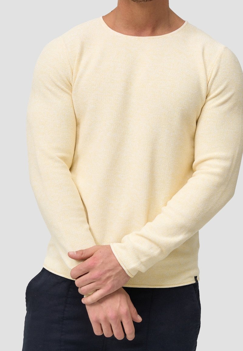 Homme portant un pull en maille texturée beige clair à manches longues et un pantalon foncé, debout les mains jointes à la taille.