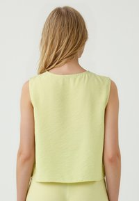 Una donna con i capelli lunghi indossa un top senza maniche di un giallo chiaro, girata di spalle contro uno sfondo semplice, mostrando il retro del vestito e la scollatura.