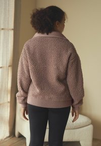 Hnědý texturovaný oversize svetr s žebrovaným límcem a manžetami, kombinovaný s černými legínami. Měkký, plyšový materiál a volný střih.