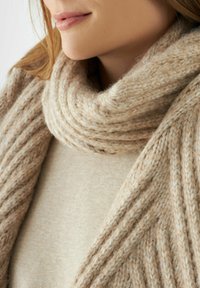 Close-up van een persoon die een beige gebreide sjaal en een bijpassende trui draagt, waarbij de zachte, dikke textuur van de wol wordt benadrukt.