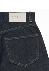 Jean en denim bleu foncé montrant la poche arrière et un patch en cuir blanc avec des caractères japonais sur la ceinture.