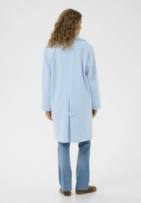 Cappotto azzurro chiaro realizzato in tessuto morbido, con una fessura sul retro chiusa da un bottone, e maniche lunghe. Abbinato a jeans azzurri chiari e scarpe marroni.