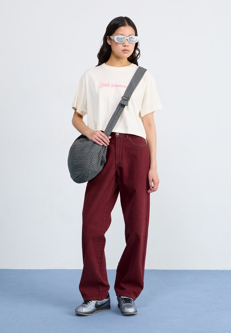 Camiseta recortada de un color beige claro con texto rosa, pantalones de pierna ancha en color burdeos, bolsa mensajera a cuadros grises y zapatillas plateadas con acentos negros.