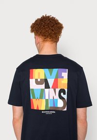 Laivastonsininen lyhythihainen t-paita, jossa on monivärinen grafiikka takana ja jossa lukee isoin kirjaimin "LOVE WINS".