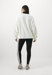 Sweater branco fluffy com um colarinho preto, combinado com leggings pretas com riscas brancas nas laterais, e sapatilhas cinzas com sola de madeira.