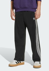 Pantalons de sport noirs avec trois rayures blanches sur les côtés, fabriqués en tissu lisse. Portés avec des baskets blanches et noires dotées de semelles en caoutchouc.