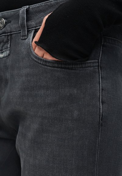 Main dans la poche avant d'un jean gris foncé, portant un haut noir à manches longues avec trou pour le pouce.