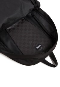 Intérieur d'un sac à dos noir montrant un revêtement à motif à carreaux, une étiquette portant le nom "Vans" et une fermeture éclair le long du bord supérieur. Détails en tissu texturé visibles.