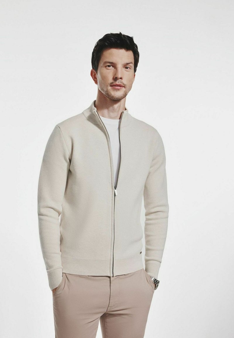 Jasno-beżowy sweter na zamek w miękkiej dzianinie, z kołnierzem stojącym i ściągaczami, noszony na białej koszuli.