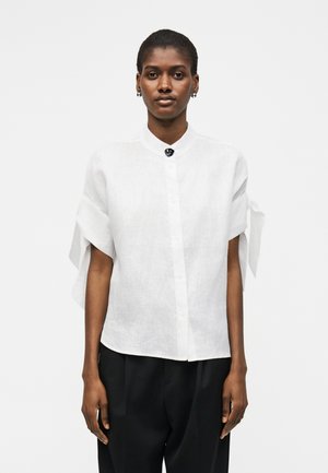 Frau trägt ein weißes kurzärmeliges Hemd mit Knöpfen und gebundenen Ärmeldetails sowie schwarze Hose, steht vor einem einfarbigen hellen Hintergrund.