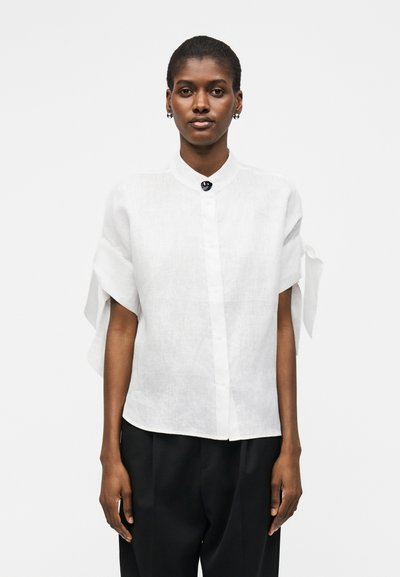 Femme portant une chemise blanche à manches courtes boutonnée avec des détails noués aux manches et un pantalon noir, debout sur un fond clair uni.