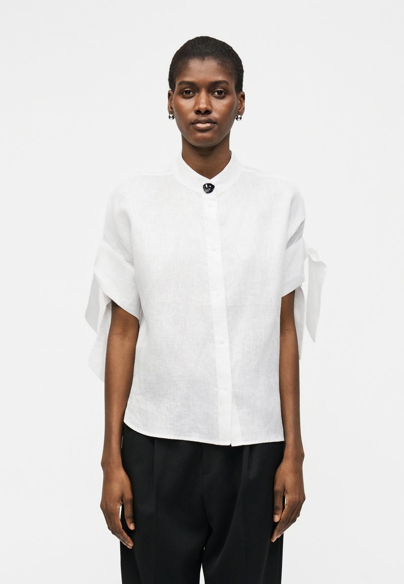 Γυναίκα που φοράει λευκό πουκάμισο με κοντά μανίκια, κουμπιά και δέσιμο στα μανίκια και μαύρο παντελόνι, στέκεται μπροστά σε απλό φωτεινό φόντο.