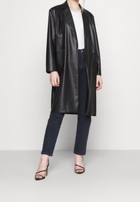 Abrigo largo de piel sintética negra con un corte holgado, solapas muescadas y bolsillos laterales, combinado con jeans oscuros y sandalias de tacón con tiras.