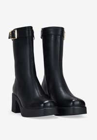 Botas de tobillo de cuero negro con tacón cuadrado, cremallera lateral y un acento de hebilla dorada en la parte superior. Textura suave y diseño aerodinámico.