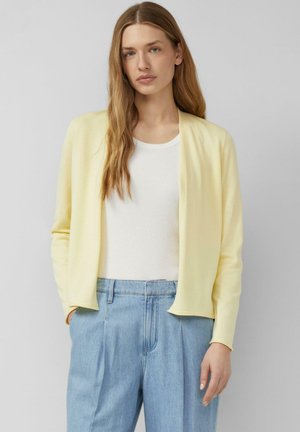 Junge Frau mit langen hellbraunen Haaren, die eine blassgelbe offene Strickjacke, ein weißes Top und hellblaue hochtaillierte Jeans trägt, steht vor einem grauen Hintergrund.
