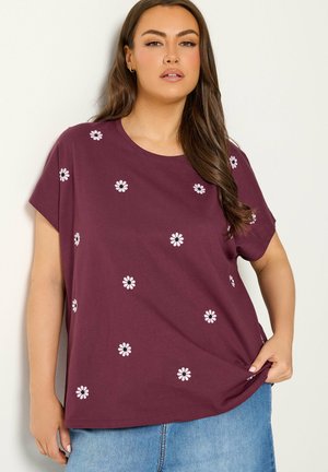 Kvinde med langt brunt hår iført en lilla T-shirt med hvide tusindfrydprint, kombineret med lyseblå jeans, stående mod en ensfarvet baggrund.