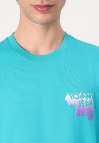 Tyrkysové bavlněné tričko s kulatým výstřihem. Obsahuje potisk s logem v bílé a fialové barvě s nápisem "TOMMY JEANS" a "NEW YORK CITY".