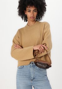 Maglione beige a maglia con scollatura a girocollo rigata e spalle scese, abbinato a jeans in denim chiaro. Borsa marrone tenuta con una mano.