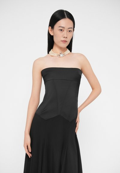 Mladá žena v čiernych šatách bez ramienok a perlovom náhrdelníku s príveskom v tvare hviezdy, stojaca pred jednoduchým bielym pozadím.