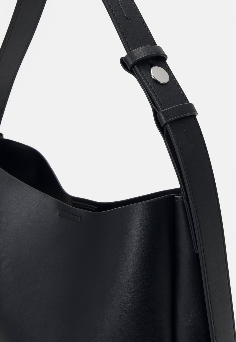 Borsa tote in pelle nera con una texture liscia. Presenta un'ampia apertura e una tracolla cucita con un dettaglio di bottone a scatto argentato.