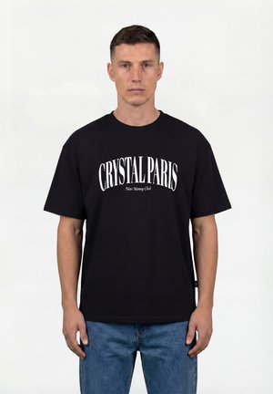 Man draagt een zwart T-shirt met witte tekst "CRYSTAL PARIS New Money Club" en een blauwe spijkerbroek tegen een effen witte achtergrond.