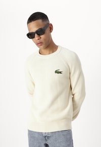 Cremfarbener, gerippter Strickpullover mit Rundhalsausschnitt, mit einem grünen Krokodil-Logo auf der linken Seite. Modell trägt schwarze, kantige Sonnenbrillen.