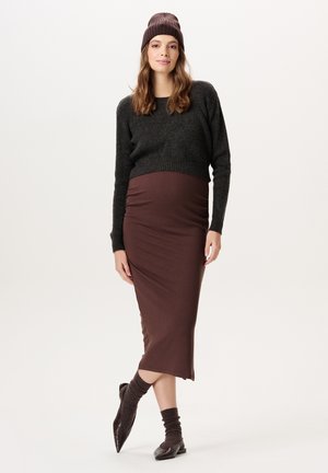 Vrouw die een donkergrijze trui, een bruine midirok, bijpassende sokken, schoenen en een gebreide muts draagt, staande met één been gekruist voor zich.