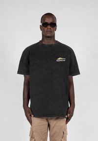 Svart oversized T-skjorte med brystgrafikk i hvitt og gull. Laget av bomull med en falmet tekstur. Kombinert med brune cargo-bukser.