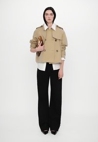 Sacou bej cu două rânduri de nasturi și epoleți, purtat cu o cămașă albă și pantaloni negri largi. Modelul ține o poșetă cu un model.