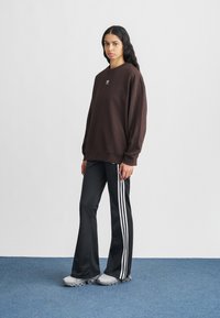 Καφέ φαρδύ φούτερ με μικρό λογότυπο, συνδυασμένο με μαύρο φαρδύ παντελόνι με λευκές κάθετες ρίγες στο πλάι και γκρι αθλητικά παπούτσια.