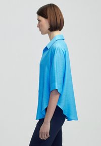 Svetle modrá košile s krátkým rukávem a límečkem, se volným střihem a mírně delším zadním dílem. Vyrobena z lehké, texturované látky.