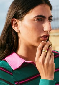 Green sweater with pink collar and stripes, gold hoop earring, gold ring on hand, natural skin texture, soft lighting.