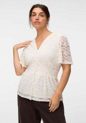 Femme aux cheveux foncés portant un chemisier boutonné en dentelle blanche à manches courtes et un pantalon sombre sur un fond clair uni.