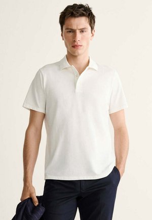 Junger Mann trägt ein weißes kurzärmeliges Polohemd und dunkle Hose, hält in einer Hand eine dunkle Jacke.