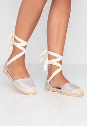Silberne Espadrilleschuhe mit gewebter Textur, beiger Jutesohle und kreuzweise verlaufenden elfenbeinfarbenen Schnürsenkeln um den Knöchel.