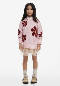 Rosa Strickpullover mit bordeauxroten Blumenmustern, übereinander getragen mit einem transparenten beigen Rock, kombiniert mit beigen Slipper-Schuhen und gerippten cremigen Socken.