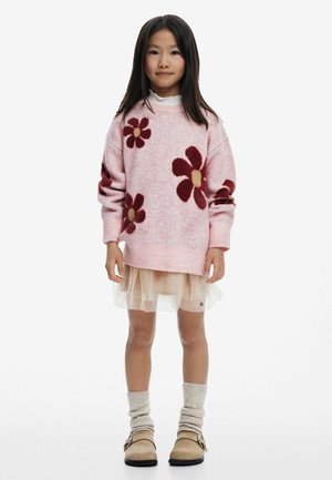 Maglione rosa a maglia con motivi floreali bordeaux, indossato sopra una gonna beige trasparente, abbinato a scarpe slip-on beige e calzini rigati color crema.