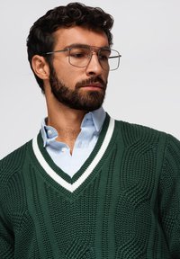 Maglione verde lavorato a maglia con scollatura a V e righe bianche, indossato sopra una camicia azzurra con colletto. Presenta un motivo testurizzato e polsini a coste.