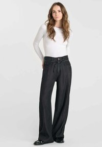 Zwarte wijde broek gemaakt van een textuur stof, met een hoge taille, voorzakken en een strikceintuur. Gecombineerd met een witte top met lange mouwen.