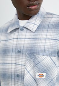 Camicia di flanella a quadri blu e bianca con colletto, due bottoni e una tasca sul petto con un'etichetta di marca. Tessuto morbido.