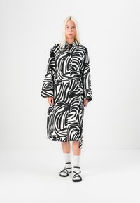 Rochie cu un model alb-negru, cu curea, mâneci lungi și buzunare. Purtați cu șosete albe și sandale negre, pe un fundal simplu.