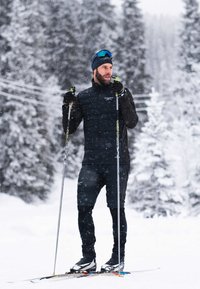 Man in zwarte wintersportkleding staand op langlaufski's in een besneeuwd bos, met skistokken in de hand tijdens vallende sneeuw.