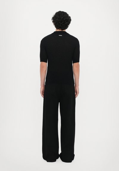 Schwarzes kurzärmliges Strickpolohemd, enganliegendes Design mit gerippter Textur, getragen mit schwarzen weiten Hosen, mit einem Logo am hinteren Kragen.