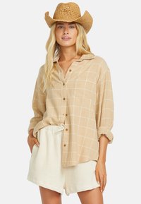 Beige kostkovaná košile na knoflíky s dlouhými rukávy a jednou náprsní kapsou, kombinovaná s texturovanými bílými šortkami. Vyrobena z lehkého materiálu.