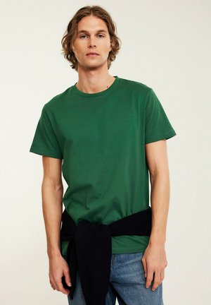 Młody mężczyzna o falowanych włosach, ubrany w zielony t-shirt i niebieskie dżinsy, z czarnym swetrem zawiązanym wokół talii, stojący na jednolitym tle.