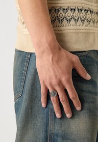 Anel de metal com um design de bússola, usado numa mão apoiada contra o denim. Vestuário de malha bege com padrões texturizados visíveis acima da cintura.