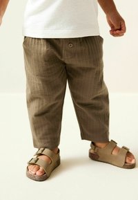 Brown textured trousers with a loose fit, featuring a button closure. Paired with brown sandals with two adjustable straps and a cork sole.